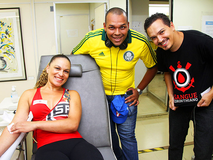 来自SobreVivência的照片-当女子献血时，两男一女穿着不同的足球队球衣微笑着